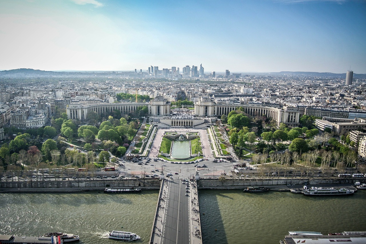 Bridge across a river to a majestic old city with park and palatial buildings, skyscrappers in the distance.